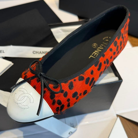 A Chanel ballet flat shoe with a vibrant orange and black leopard print upper, a white leather toe cap with an embossed Chanel logo, and a black ribbon bow, displayed on its black box packaging.