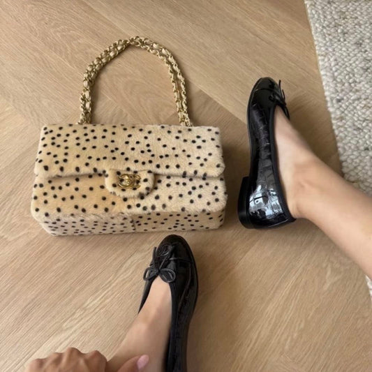A person wearing black patent leather ballet flats with bow details, shown from above on a light wood floor next to a beige and black polka dot handbag with a gold chain strap.