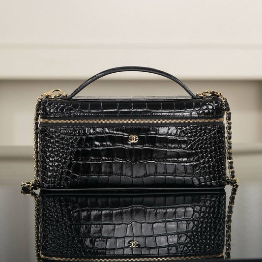 A black crocodile-embossed leather handbag with gold-tone chain strap and hardware, displayed on a reflective surface against a soft beige background.