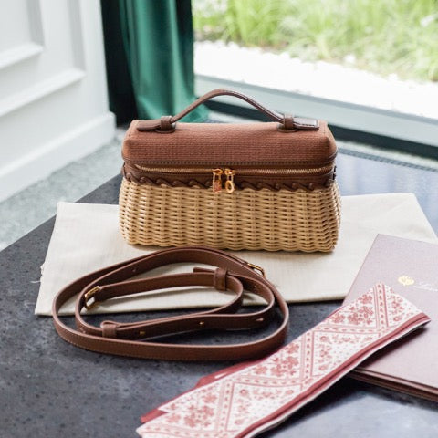 A woven rectangular cosmetic case with a brown top and handle, a matching brown shoulder strap, and a gold zipper, displayed on a dark tabletop with a cream dust bag and patterned accessories nearby. No person or animal is present, but the real interior setting and styled props make this a Lifestyle image rather than a pure studio Product/Packaging shot.