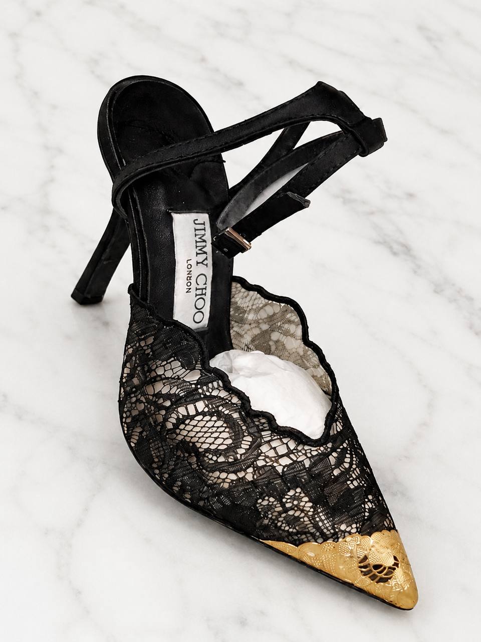 Black lace high-heeled shoe with gold tip on a white marble surface, featuring Jimmy Choo branding.